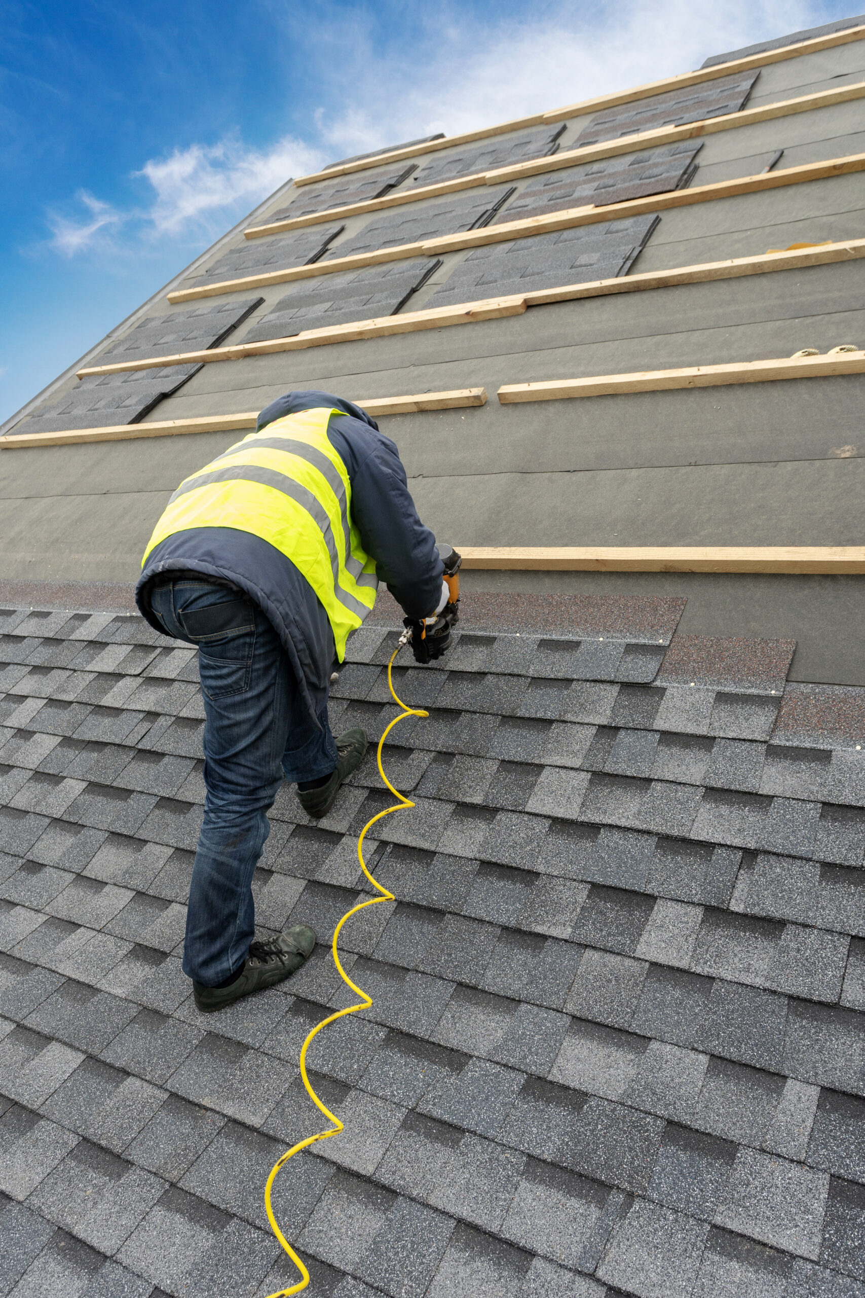 worker in uniform work wear using air or pneumatic nail gun and installing asphalt or bitumen tile on top of the roof under construction